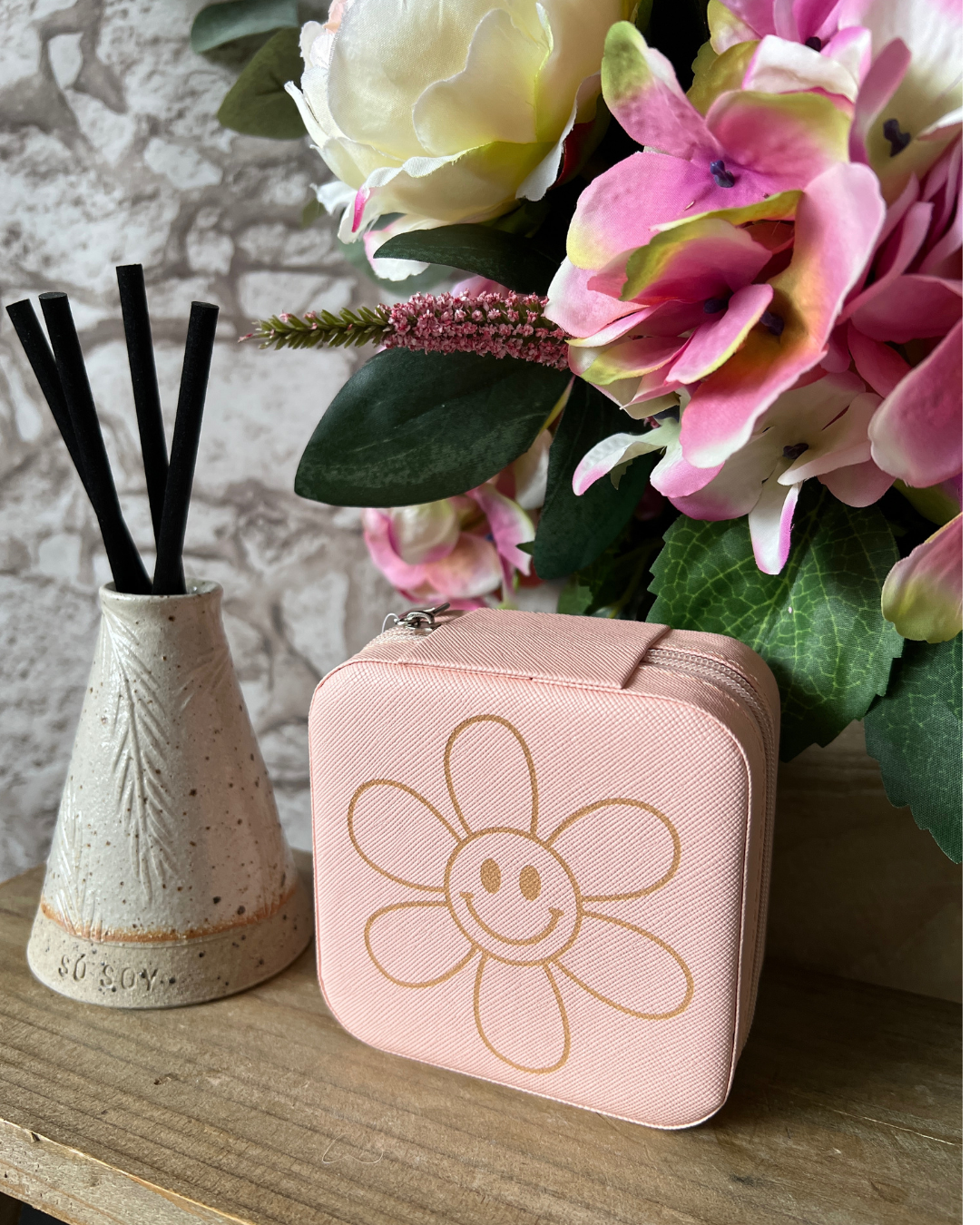 Pink case with flower design next to a decorative vase with black sticks on a wooden surface.