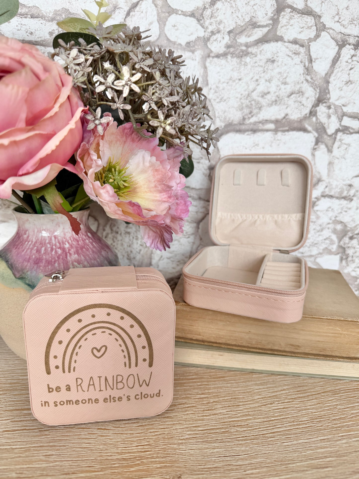 Small pink jewelry box with rainbow design and text "be a rainbow in someone else's cloud", next to an open jewelry case on a wooden surface with flowers in the background.