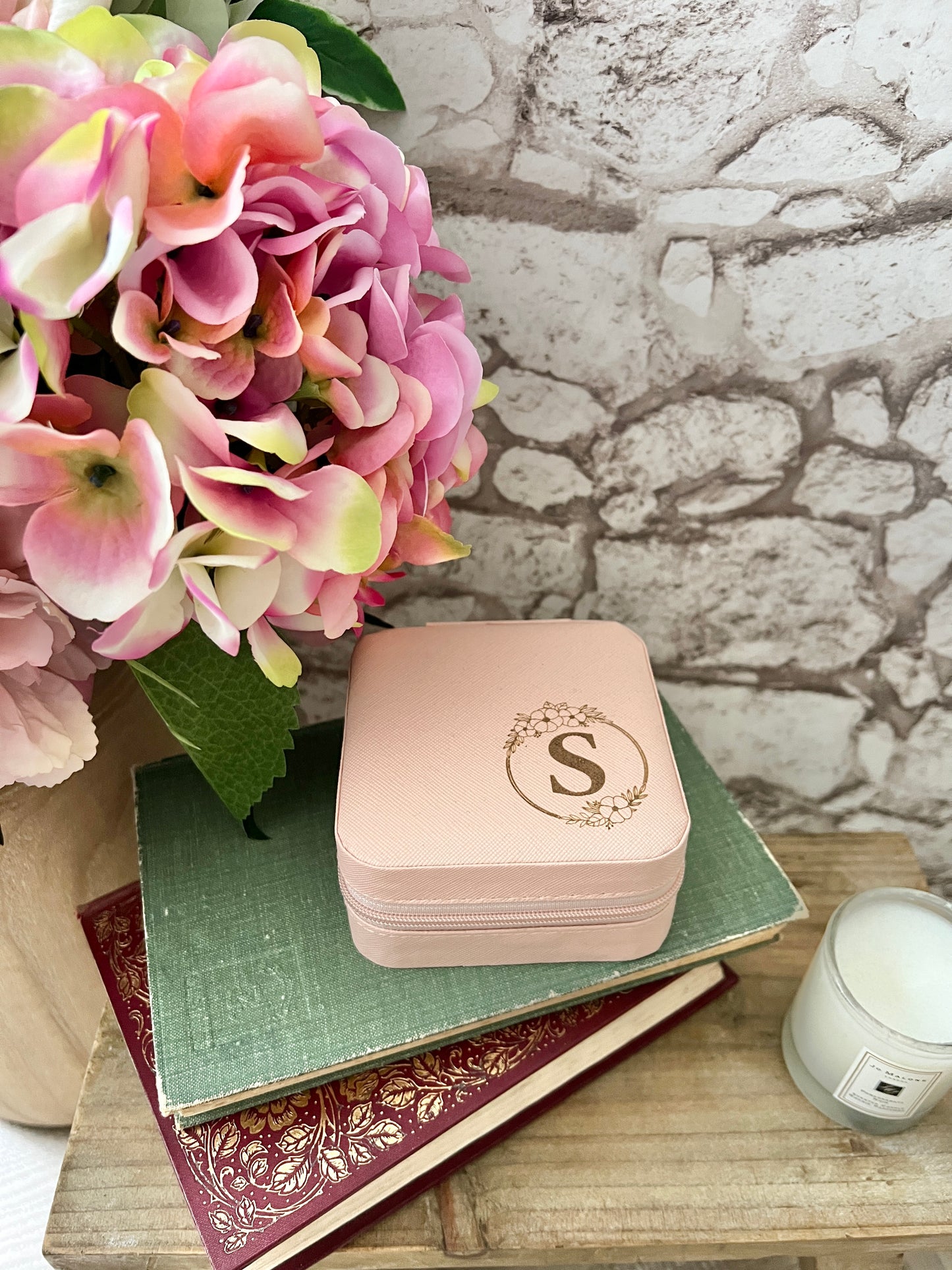 A pink personalised jewellery box with a floral design and a initial 'S', placed on top of a green book, next to a brick wall and a pink floral decoration.
