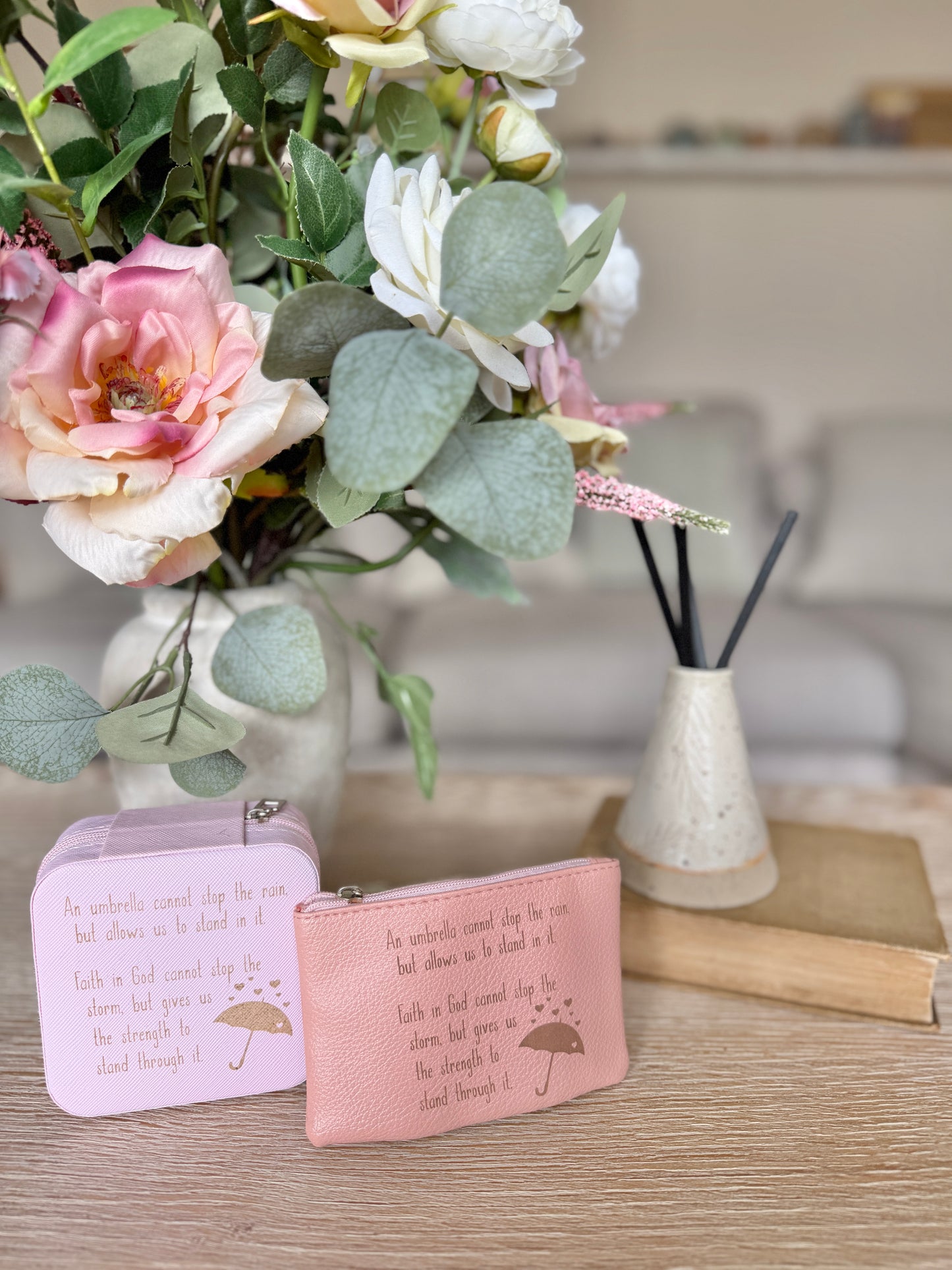 A pink jewellery box alongside pink coin purse with an umbrella image and the text "An umbrella cannot stop the rain but allows us to stand in it. Faith in God cannot stop the storm, but gives us the strength to stand through it" on a surface with flowers and a diffuser in the background.