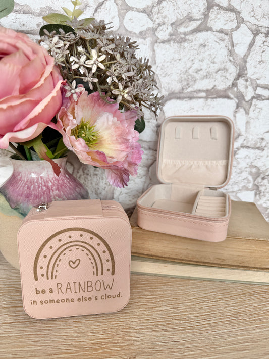 Small pink jewelry box with rainbow design and text "be a rainbow in someone else's cloud", next to an open jewelry case on a wooden surface with flowers in the background.