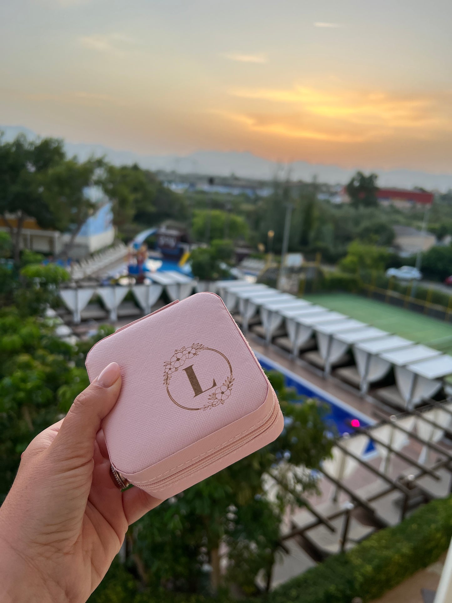 Pink jewellery box with circle floral detail with letter L in the middle. Holiday destination in the background.