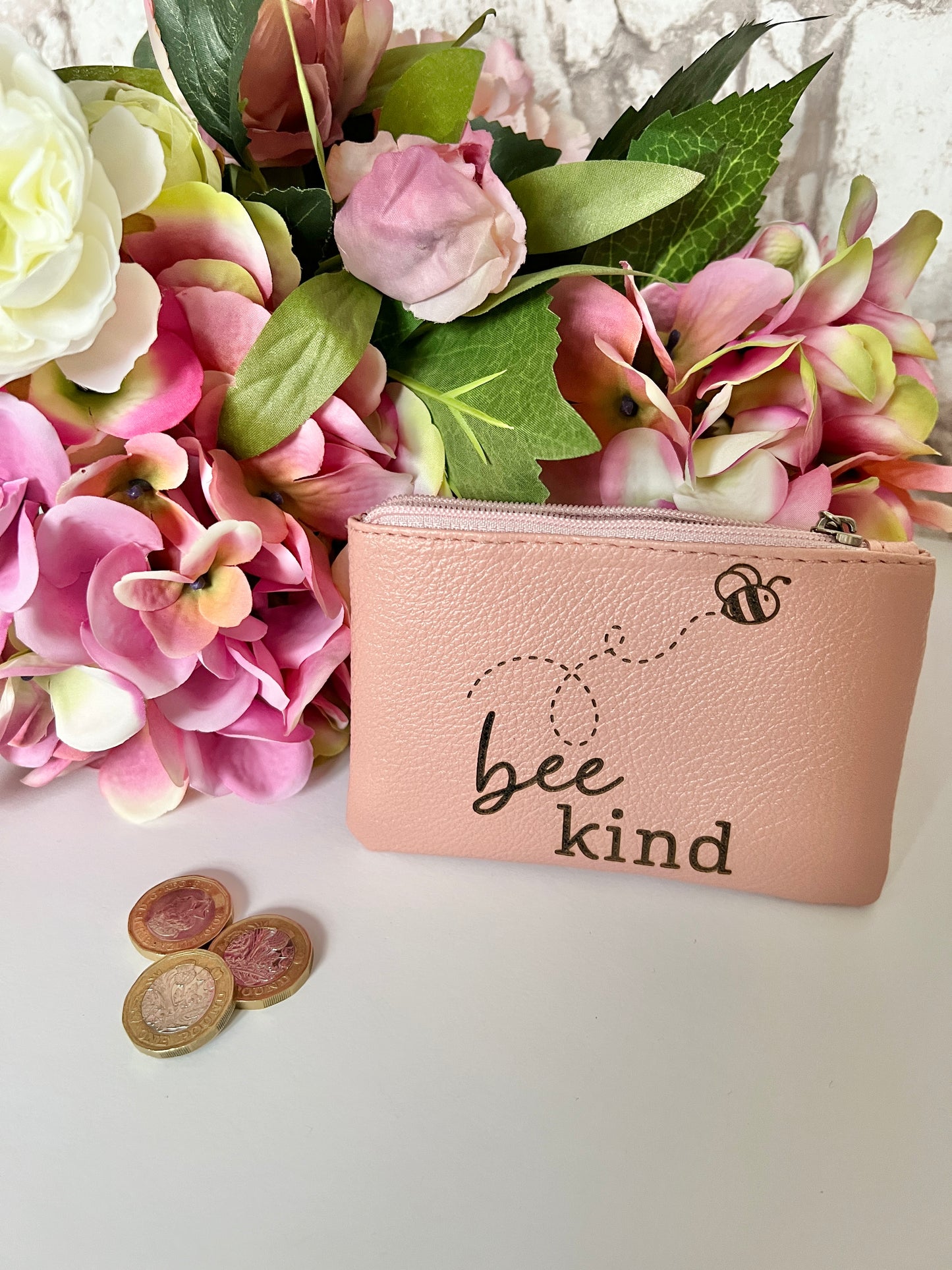 A pink coin purse with image of a bee and the text "bee kind" with flowers in the background