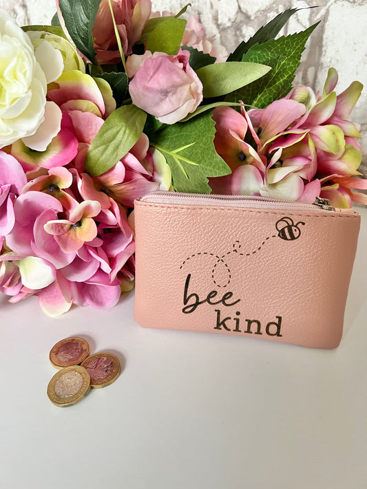 A pink coin purse with image of a bee and the text "bee kind" with flowers in the background