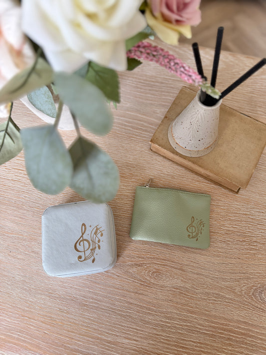 A light blue jewellery box alongside a sage green coin purse with treble clef and music notes, on a wooden surface with flowers and a diffuser in the background.